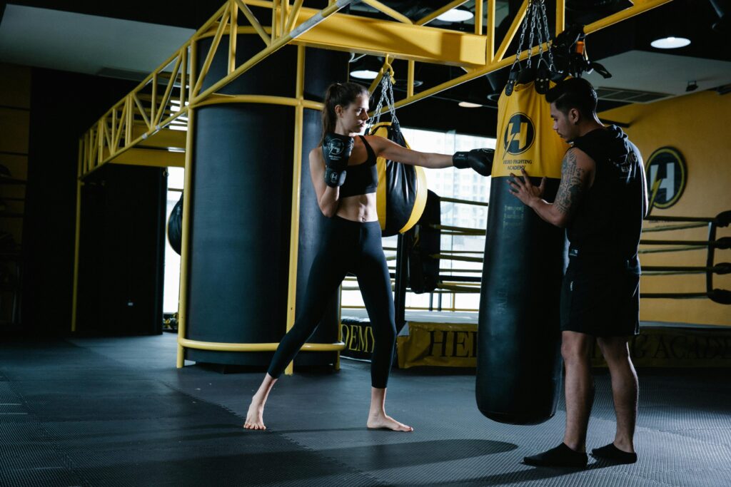 Full body of sportive female fighter punching boxing bag during workout with tattooed personal Latin American instructor in modern gym